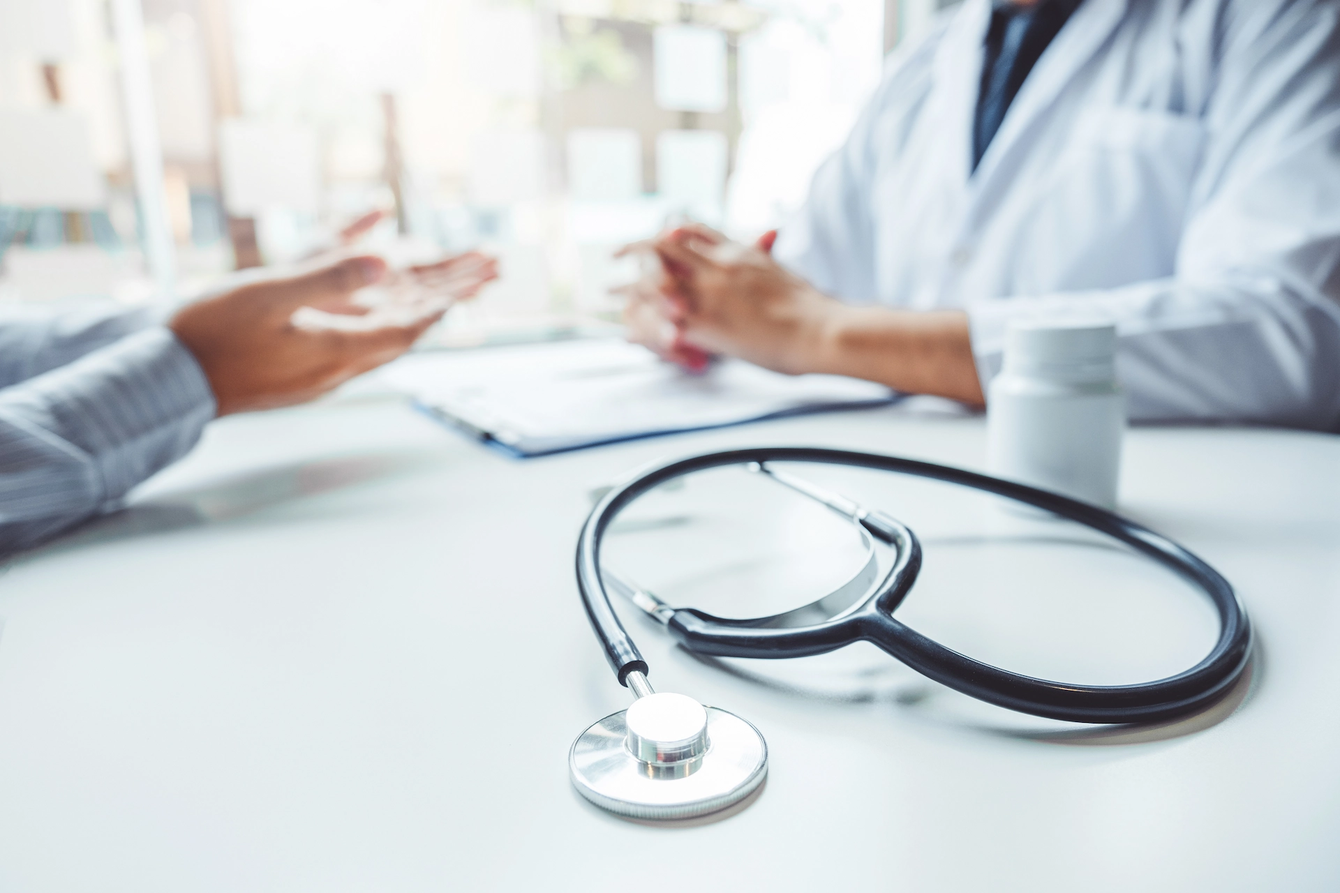 Stethoscope resting on a desk in the foreground with a doctor and patient consulting in the blurred background
